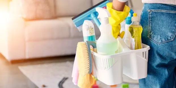 A woman facing her sofa while wearing cleaning gloves and holding a cleaning caddy. 