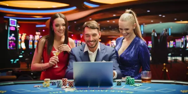 A man and two women smiling at a laptop screen while sitting at a poker table. 