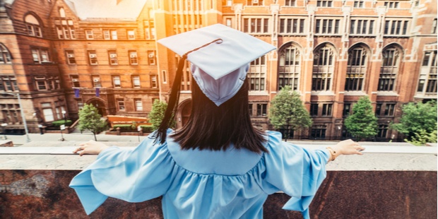 A college graduate, wearing a blue robe and matching hat, standing on the rooftop of a building and looking out for her future. 