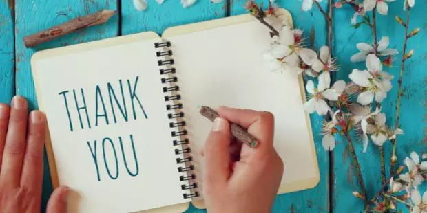 A woman writing "thank you" in a notebook, placed on a wooden table near cherry blossom flowers. 