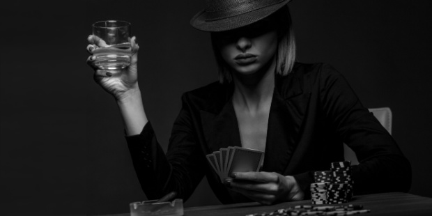 A black and white photo of a woman sitting at the poker tables, playing cards, and holding a glass. 