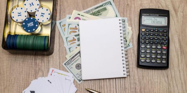 A notebook and a calculator appear next to poker chips and stacks of cash. 
