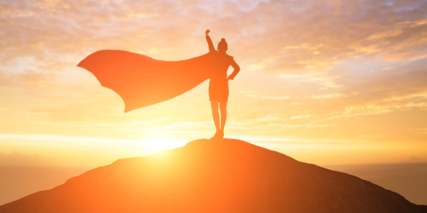 A woman wearing a cape holding a Superwoman pose on top of a hill. 