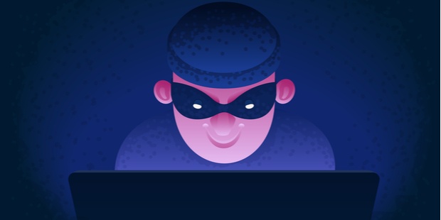 A thief in mask and dark clothes sits behind a computer. 